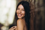 A woman with long dark hair is smiling warmly. She is leaning against a textured surface, with soft lighting creating a gentle ambiance.