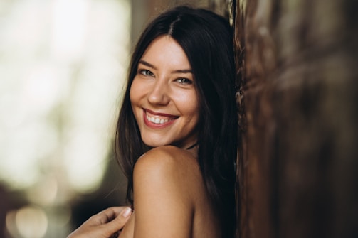 A woman with long dark hair is smiling warmly. She is leaning against a textured surface, with soft lighting creating a gentle ambiance.
