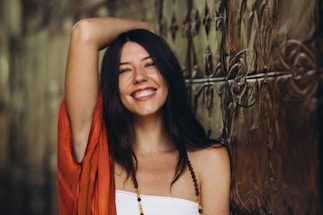 A woman with long dark hair and a bright smile poses with one arm raised, wearing an orange garment and a beaded necklace. She leans against an ornate metallic wall with an intricate pattern.