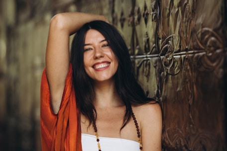 A woman with long dark hair and a bright smile poses with one arm raised, wearing an orange garment and a beaded necklace. She leans against an ornate metallic wall with an intricate pattern.