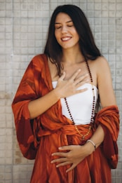 A woman with long dark hair is wearing a flowing orange garment and a beaded necklace. She has a relaxed and content expression, with her eyes closed and hands placed over her chest and stomach. The background features a tiled wall.