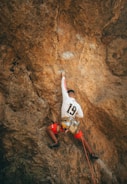 A person engaged in outdoor rock climbing, wearing red shorts and a white shirt with the number 19. They are equipped with a harness and rope while navigating a large, rugged rock face. The rock appears to be a tan or light brown in color.