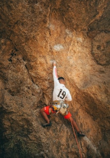 A person engaged in outdoor rock climbing, wearing red shorts and a white shirt with the number 19. They are equipped with a harness and rope while navigating a large, rugged rock face. The rock appears to be a tan or light brown in color.