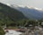 A scenic landscape featuring a lush forest and a river with a sturdy bridge crossing over it. On the opposite side of the river, several small buildings and tents are scattered amidst the greenery. Snow-capped mountains form a majestic backdrop, with clouds just starting to cover parts of the peaks.
