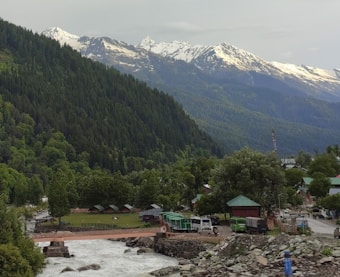 A scenic landscape featuring a lush forest and a river with a sturdy bridge crossing over it. On the opposite side of the river, several small buildings and tents are scattered amidst the greenery. Snow-capped mountains form a majestic backdrop, with clouds just starting to cover parts of the peaks.