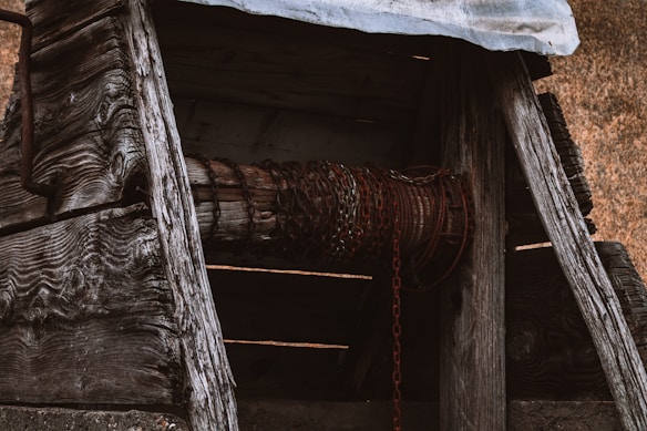 A weathered wooden structure with intricate grain patterns is featured. Chains are wrapped around a wooden cylinder, giving a rustic, old-world appearance. The structure appears to be part of an old well or winch, with a rough metallic roof and aged construction materials.