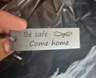 A metal keychain is held between two fingers. It features the engraved text 'Be safe Come home' and an elegant decorative scroll design.
