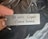 A metal keychain is held between two fingers. It features the engraved text 'Be safe Come home' and an elegant decorative scroll design.
