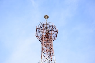 A tall, metal communication tower stretches upwards into a clear blue sky. The structure is composed of a lattice framework and has an antenna-like apparatus at the top.