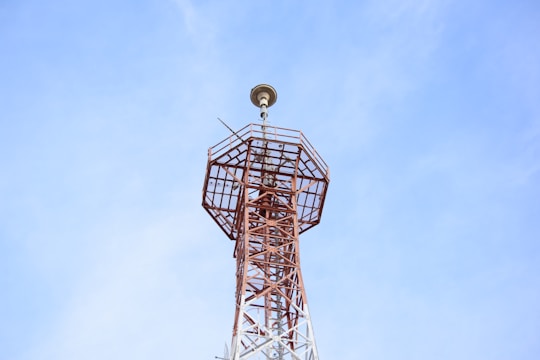 A tall, metal communication tower stretches upwards into a clear blue sky. The structure is composed of a lattice framework and has an antenna-like apparatus at the top.