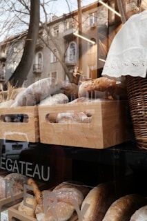 A bakery window display showcasing various fresh loaves of bread arranged in wooden crates. The reflection of trees and buildings is visible on the glass, and there is a basket with a white cloth draped over it.