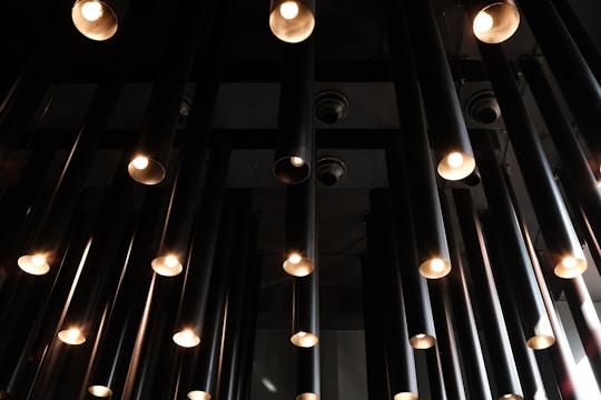 A collection of cylindrical light fixtures hangs from the ceiling, each with a warm glowing bulb. The fixtures are uniformly arranged, creating a geometric pattern against a dark background.