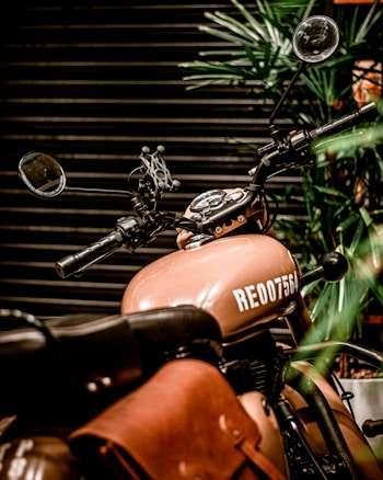 A vintage-style motorcycle with a beige tank and saddlebag, surrounded by potted greenery. The bike features classic handlebars, side mirrors, and a partially visible leather seat, all set against a dark, horizontal-striped background.