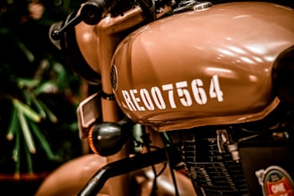 A close-up view of a tan-colored motorcycle, focusing on the fuel tank with the number RE-007564 painted in white. The badge or logo near the tank is visible, as well as parts of the engine and handlebar. The background is slightly blurred, featuring some greenery.