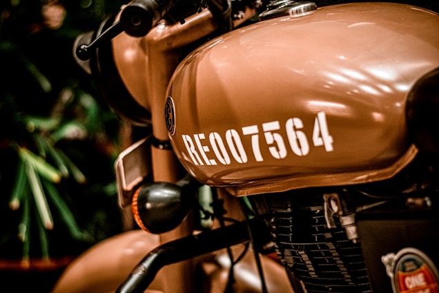 A close-up view of a tan-colored motorcycle, focusing on the fuel tank with the number RE-007564 painted in white. The badge or logo near the tank is visible, as well as parts of the engine and handlebar. The background is slightly blurred, featuring some greenery.