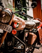 A close-up view of a vintage-style motorcycle with a brown body. The motorcycle features a round headlamp, a prominent fuel tank with decals, and a black leather seat. The license plate is visible with the number 'B5788'. The motorcycle is equipped with leather saddlebags and is positioned indoors against a blurred background.
