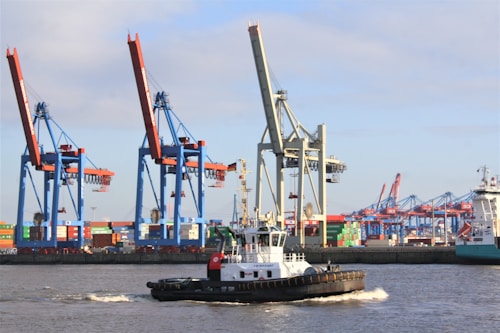 A busy harbor scene features large cranes with red and blue structures standing tall in the background, which appear to be loading or unloading colorful shipping containers. A small tugboat is navigating the choppy waters in the foreground. The sky is clear with a few clouds, adding to the industrial vibe of the location.