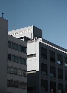 A modern office building with a minimalist design featuring large rectangular windows, flat walls, and a simplistic facade. The building is bathed in natural light under a clear blue sky, highlighting its angular architecture.