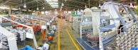 A large industrial factory setting with rows of textile machinery and spools of yarn. The machines are organized in parallel lines, and there are various structures supporting the production process. Overhead lights illuminate the space, and the floor has a tiled pattern. The presence of vibrant colors like orange and blue adds contrast to the metallic and neutral tones of the machinery.