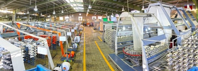 A large industrial factory setting with rows of textile machinery and spools of yarn. The machines are organized in parallel lines, and there are various structures supporting the production process. Overhead lights illuminate the space, and the floor has a tiled pattern. The presence of vibrant colors like orange and blue adds contrast to the metallic and neutral tones of the machinery.