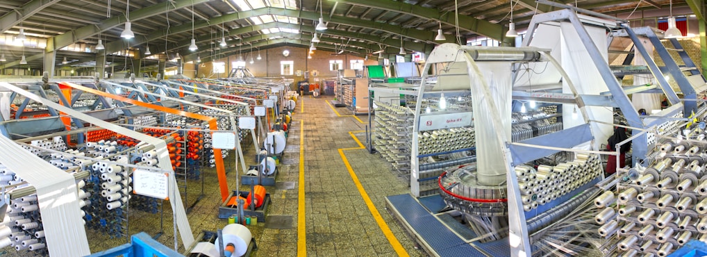 A large industrial factory setting with rows of textile machinery and spools of yarn. The machines are organized in parallel lines, and there are various structures supporting the production process. Overhead lights illuminate the space, and the floor has a tiled pattern. The presence of vibrant colors like orange and blue adds contrast to the metallic and neutral tones of the machinery.