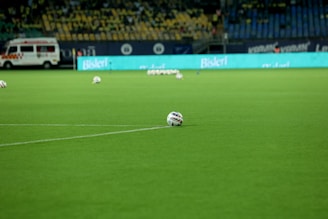 A soccer field with bright green grass and several soccer balls scattered across the turf. An ambulance is parked at the edge of the field in the background, alongside a stadium seating area filled with empty yellow and blue seats. An illuminated advertisement board displays a bright aqua-colored sign.