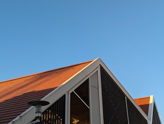 A modern building with a triangular roof, featuring reddish-brown tiles and a metallic structure. The clear blue sky contrasts with the warm tones of the roof. The building has vertical slats and a visible outdoor heater or light fixture.
