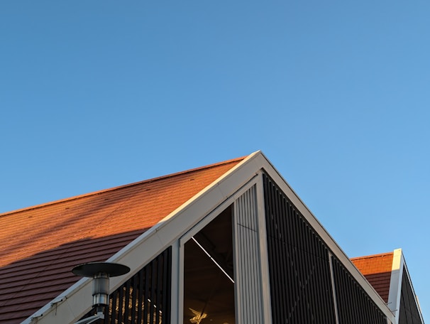 A modern building with a triangular roof, featuring reddish-brown tiles and a metallic structure. The clear blue sky contrasts with the warm tones of the roof. The building has vertical slats and a visible outdoor heater or light fixture.