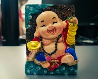 A cheerful, chubby figurine depicting a smiling, seated figure holding a string of coins in one hand and a gold ingot in the other. The figure is adorned with a red robe featuring blue and gold accents, and a black beaded necklace around its neck. The background includes decorative turquoise wave patterns and a dark textured surface, with other objects out of focus.