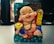 A cheerful, chubby figurine depicting a smiling, seated figure holding a string of coins in one hand and a gold ingot in the other. The figure is adorned with a red robe featuring blue and gold accents, and a black beaded necklace around its neck. The background includes decorative turquoise wave patterns and a dark textured surface, with other objects out of focus.