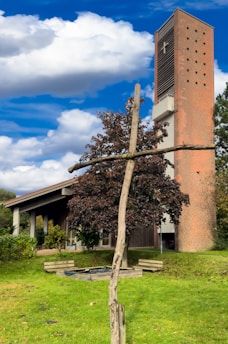 A red brick church tower with a cross stands beside a brown-leaved tree. A wooden cross structure is visible in the foreground, set against a background of vibrant green grass. The sky is bright blue with several white clouds scattered across it.