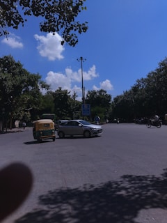A street scene with several vehicles including a green and yellow auto rickshaw, a silver car, and a bicycle. Trees with dense green foliage line the road under a bright blue sky with a few clouds. A streetlight and traffic signs are visible, and there are shadows on the road surface.