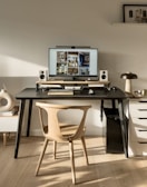 A modern workspace featuring a large computer monitor displaying interior design images and flanked by speakers on a black desk. Below the monitor, a wooden keyboard and mouse are placed. The setup includes a wooden chair and a side table with stacked books and decorative items. A small lamp and various decorative objects are arranged on the desk and adjacent surfaces.