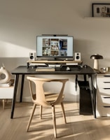 A modern workspace featuring a large computer monitor displaying interior design images and flanked by speakers on a black desk. Below the monitor, a wooden keyboard and mouse are placed. The setup includes a wooden chair and a side table with stacked books and decorative items. A small lamp and various decorative objects are arranged on the desk and adjacent surfaces.