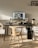 A modern workspace featuring a large computer monitor displaying interior design images and flanked by speakers on a black desk. Below the monitor, a wooden keyboard and mouse are placed. The setup includes a wooden chair and a side table with stacked books and decorative items. A small lamp and various decorative objects are arranged on the desk and adjacent surfaces.