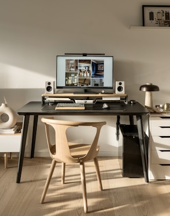 A modern workspace featuring a large computer monitor displaying interior design images and flanked by speakers on a black desk. Below the monitor, a wooden keyboard and mouse are placed. The setup includes a wooden chair and a side table with stacked books and decorative items. A small lamp and various decorative objects are arranged on the desk and adjacent surfaces.