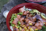 A vibrant bowl filled with fresh ingredients including cut salmon, avocado slices, edamame beans, corn, and drizzled with a dark sauce. The mixture is arranged in a red bowl on a rustic surface.