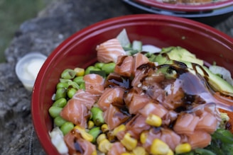 A vibrant bowl filled with fresh ingredients including cut salmon, avocado slices, edamame beans, corn, and drizzled with a dark sauce. The mixture is arranged in a red bowl on a rustic surface.