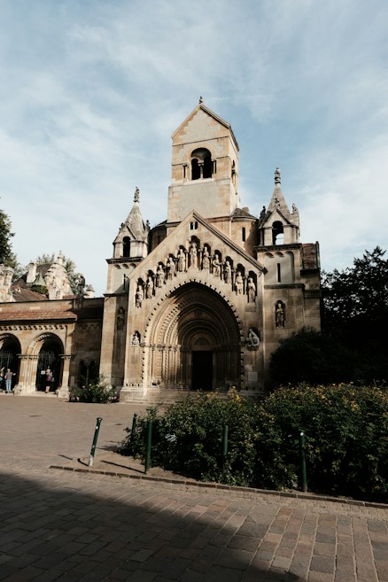 A historic, ornate church or cathedral facade with detailed stone carvings and arches. The building has a prominent bell tower, intricate arches over the entrance, and decorative statues embedded in the structure. It is set against a clear blue sky, surrounded by greenery and a cobblestone path in the foreground.