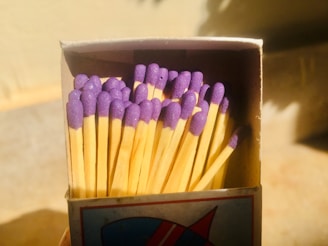 A close-up of a matchbox filled with matches. The matches have wooden sticks and purple heads. The surrounding lighting and shadows create a warm, contrasting effect.