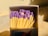 A close-up of a matchbox filled with matches. The matches have wooden sticks and purple heads. The surrounding lighting and shadows create a warm, contrasting effect.