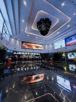 A modern cinema lobby features a high ceiling with intricate lighting designs and a large, decorative chandelier. Multiple screens displaying movie posters and information line the walls, and a concession stand is visible in the background. The glossy, reflective floor captures the ceiling's designs and advertisements.