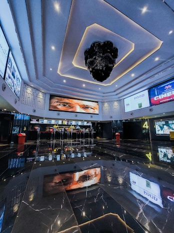 A modern cinema lobby features a high ceiling with intricate lighting designs and a large, decorative chandelier. Multiple screens displaying movie posters and information line the walls, and a concession stand is visible in the background. The glossy, reflective floor captures the ceiling's designs and advertisements.