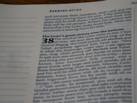 An open page from a Bible, displaying text from the book of Ezekiel, chapter 37 verse 27 and part of chapter 38. The text includes a passage about a prophecy and makes references to places and names such as Gog, Magog, Meshek, and Tubal. The page is slightly angled, showing a focus on the central column of text with italic and bold formatting for emphasis.