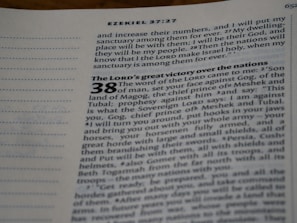 An open page from a Bible, displaying text from the book of Ezekiel, chapter 37 verse 27 and part of chapter 38. The text includes a passage about a prophecy and makes references to places and names such as Gog, Magog, Meshek, and Tubal. The page is slightly angled, showing a focus on the central column of text with italic and bold formatting for emphasis.