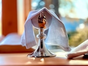A polished silver chalice is partially covered with a white cloth, placed on a wooden surface. The background is softly blurred, with hints of natural light filtering through, suggesting a serene and contemplative atmosphere.