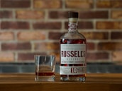 A bottle of Russell's Reserve Kentucky Straight Bourbon Whiskey is placed on a wooden surface with a glass containing a small amount of the whiskey next to it. The background features a brick wall with a slightly blurred texture.