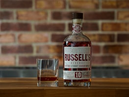 A bottle of Russell's Reserve Kentucky Straight Bourbon Whiskey is placed on a wooden surface with a glass containing a small amount of the whiskey next to it. The background features a brick wall with a slightly blurred texture.