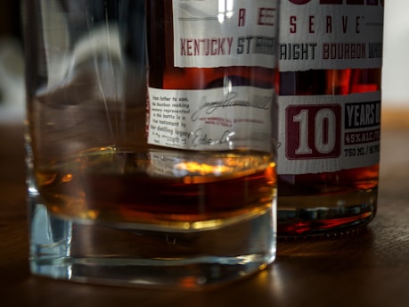 A close-up view of a glass of whiskey alongside a bottle of bourbon with visible labels detailing its contents and age. The label indicates '10 years old' and mentions the alcohol content. The scene is set on a wooden surface, and the whiskey in the glass casts a warm, amber reflection.