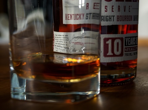 A close-up view of a glass of whiskey alongside a bottle of bourbon with visible labels detailing its contents and age. The label indicates '10 years old' and mentions the alcohol content. The scene is set on a wooden surface, and the whiskey in the glass casts a warm, amber reflection.